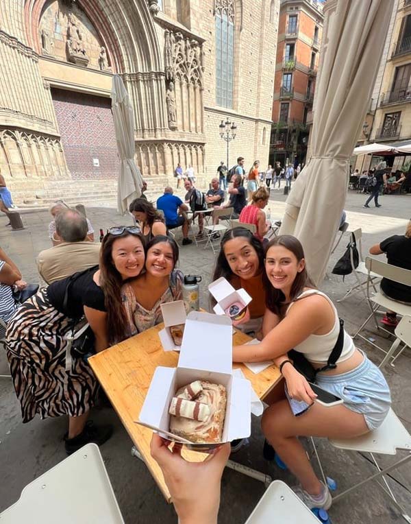 一群留学生坐在桌子旁，桌上放着一盒盒的食物