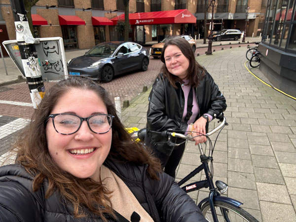 两名留学生站在城市人行道上的自行车旁，微笑着自拍