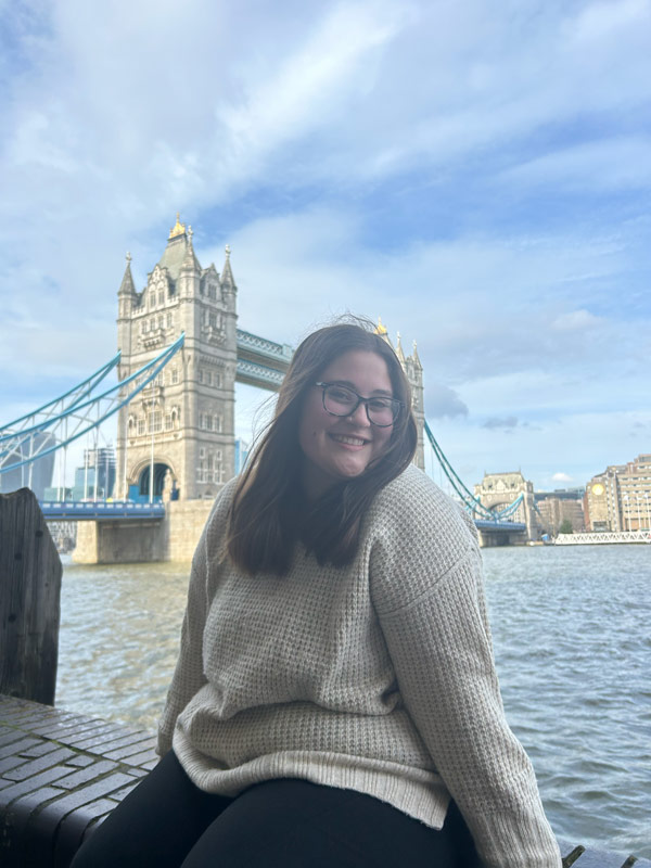 一个留学生坐在墙上，对着镜头微笑，前方是一片水域和一座大桥