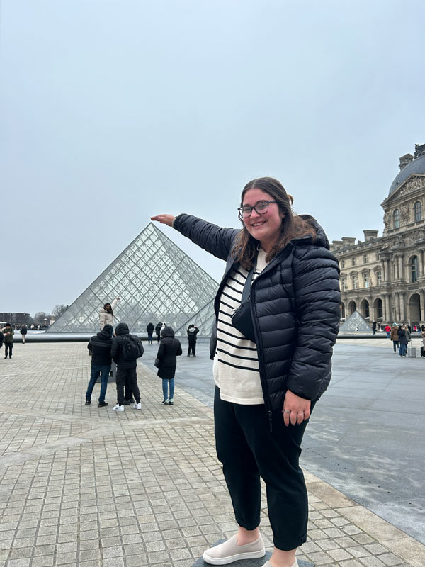 一名留学生站在卢浮宫玻璃金字塔前，对着镜头微笑