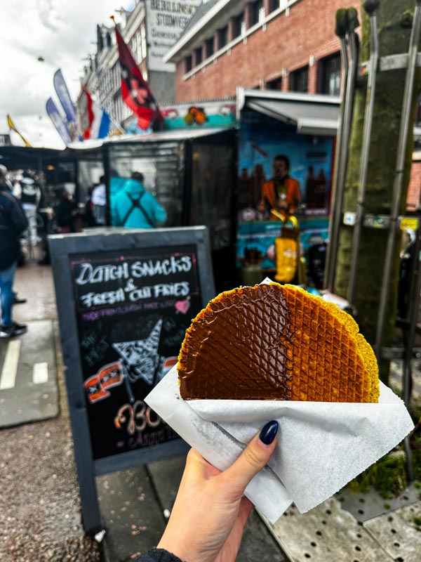 A hand holding a stroopwafel half covered in chocolate with a busy street in the background