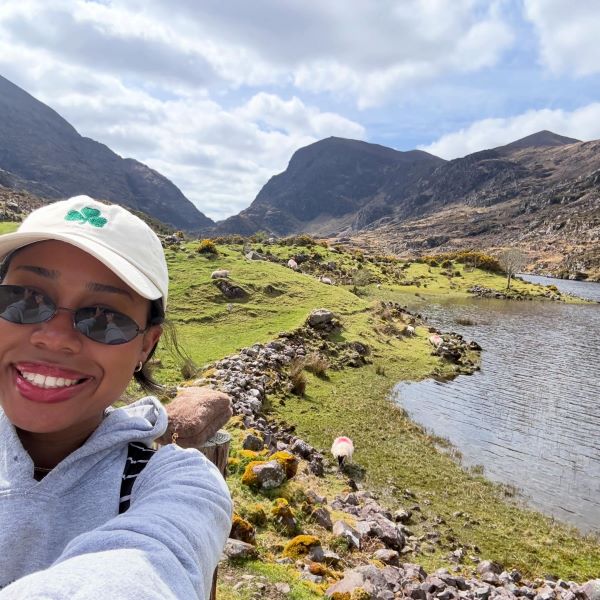 A study abroad student wearing sunglasses and a white baseball hat taking a selfie in front of an expansive green landscape