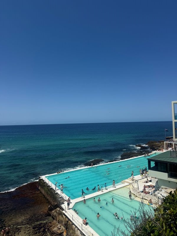 People swimming in a large clear blue pool in the foreground with the darker blue ocean in the background