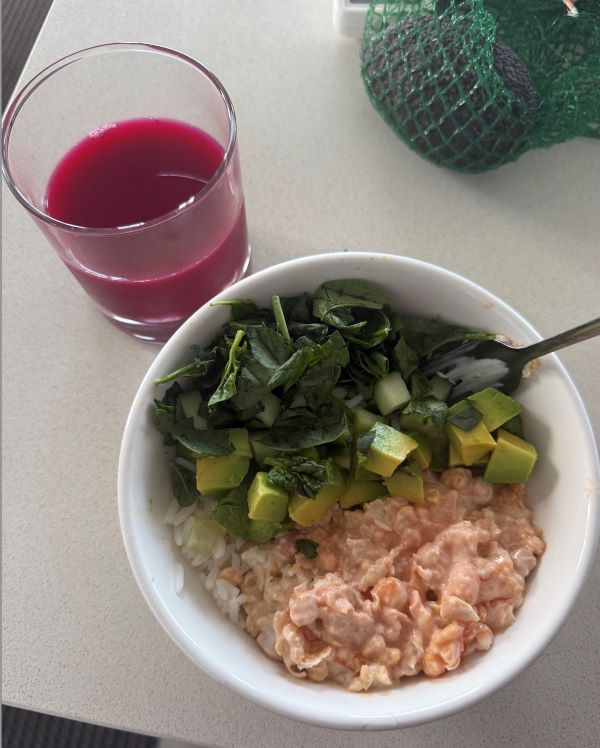 A white bowl full of mashed chickpeas, chopped avocado, and spinach leaves next to a glass of a dark pink liquid
