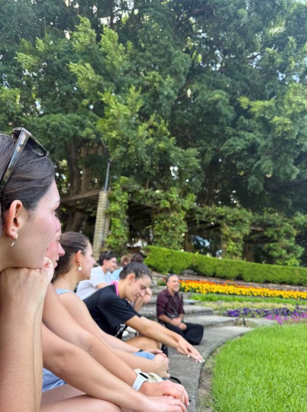 A group of study abroad students sitting outdoors to the left of the frame, with their professor at the far end of the group