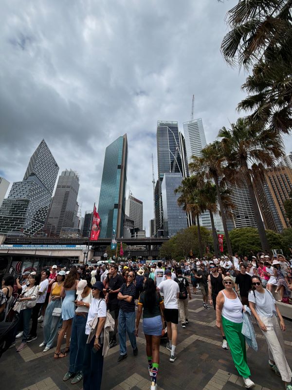 A large group of people walking in various directions outdoors with large city buildings standing tall in the background