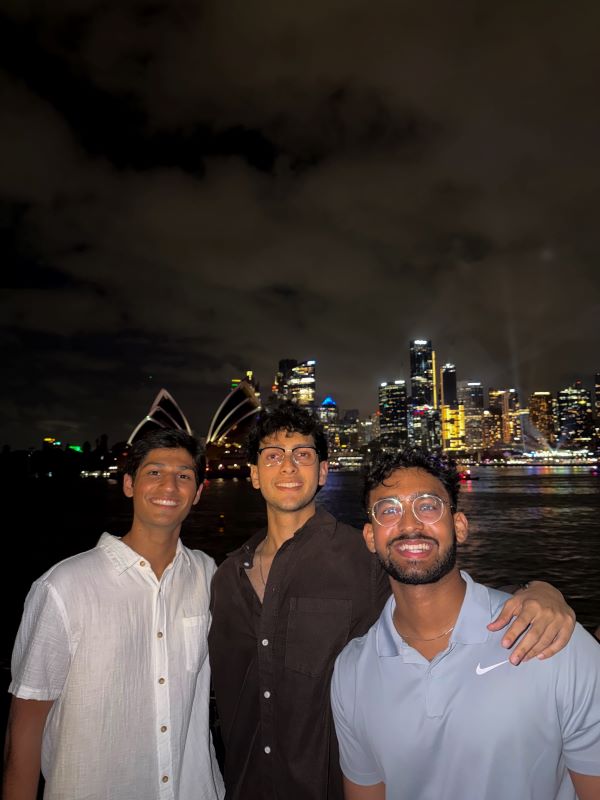 Three study abroad students smiling at the camera at nighttime with a brightly-lit city landscape behind them