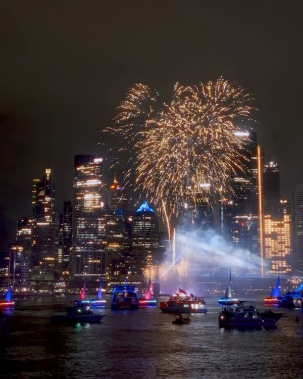 Bright explosions of fireworks seen over a city harbor at night with tall brightly-lit skyscapers in the background