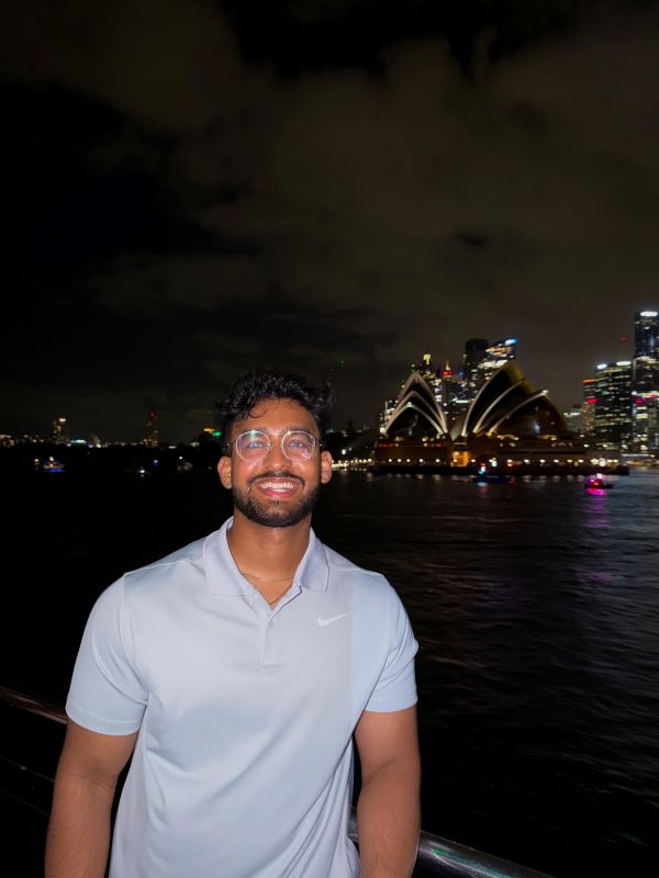 A study abroad student wearing a blue shirt smiling at the camera at night with brightly-lit city buildings in the background