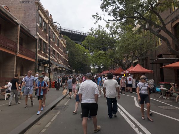 A group of people walking in various directions along a city street lined with trees and vendor's stalls