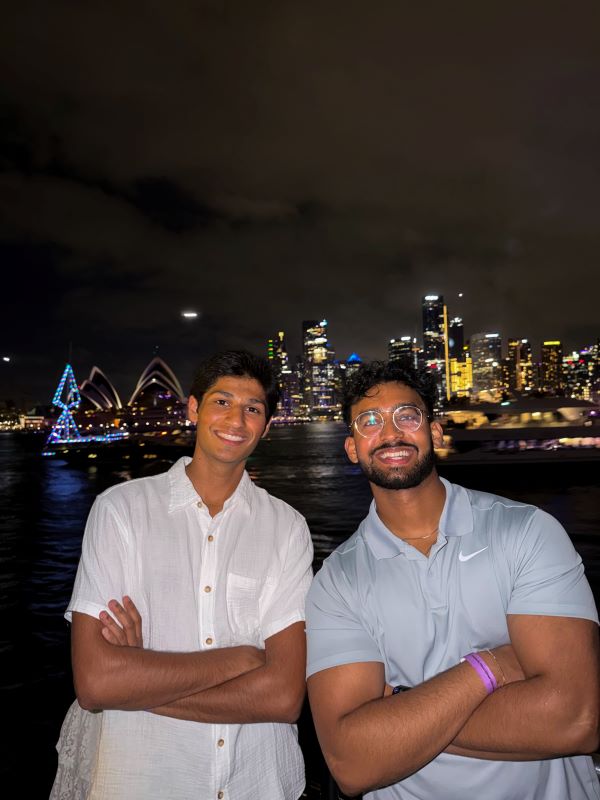 Two study abroad students smiling at the camera with their arms crossed at nighttime with city buildings lit up brightly in the distance behind them