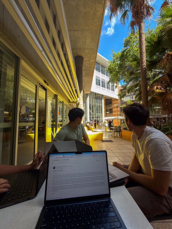 Two study abroad students seated behind a laptop on a table outdoors and looking away from the camera