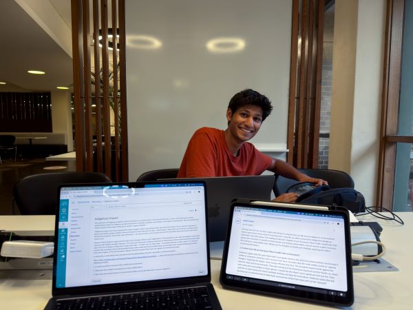 A study abroad student seated at a desk indoors with two laptops facing the viewer in front of them