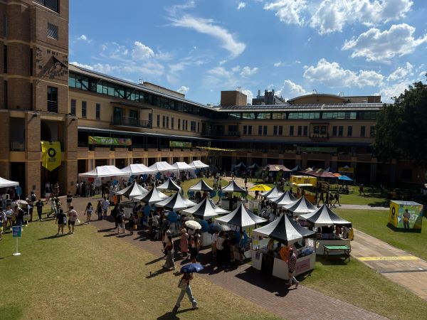 People walking around an open green space on a university campus filled with black-and-white stalls and surrounded by large brown buildings