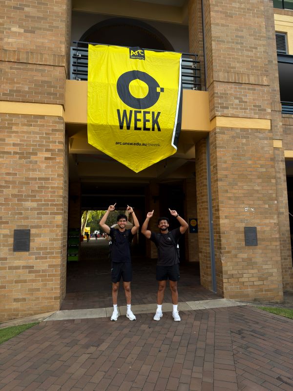 Two study abroad students smiling at the camera and standing outdoors while pointing upwards with both hands at a large yellow sign that reads