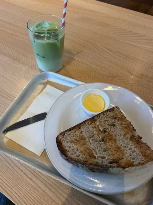 A silver tray sat on a wooden table and featuring a plate of toast with a small dish of yellow butter next to a glass of green matcha with a red-and-white striped straw