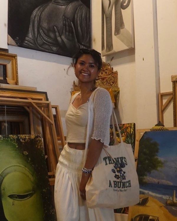 A study abroad student wearing white pants and a white shirt and holding a canvas bag on their left shoulder while smiling at the camera