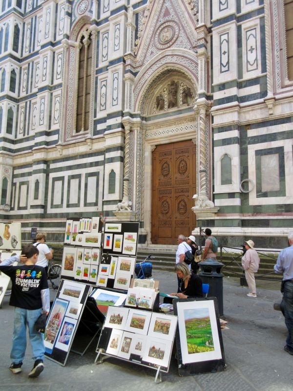 People milling around a woman seated behind a collection of art displayed on boards in an outdoor setting