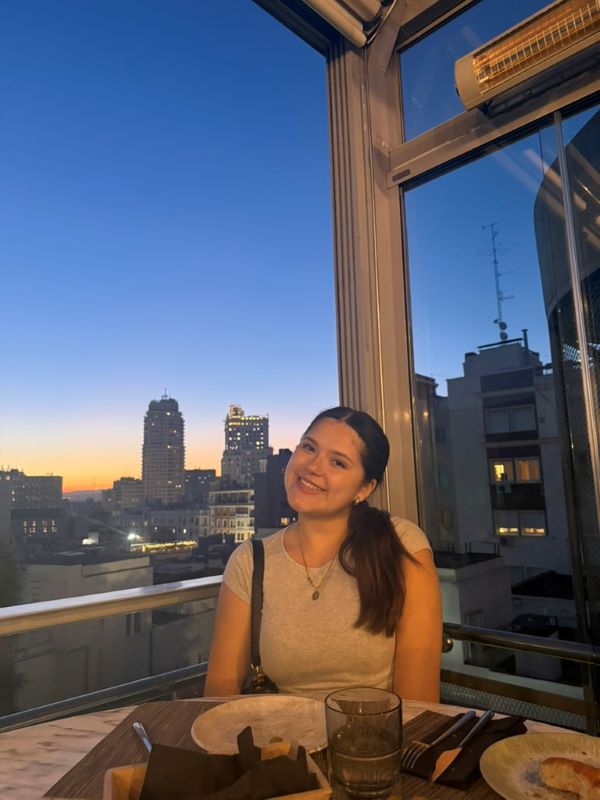A study abroad student seated at an indoor restaurant table and smiling at the camera with a city landscape seen through windows in the background