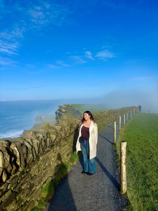 A study abroad student standing on a path outdoors with a large body of water in the distance, smiling and looking slightly away from the camera