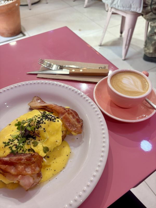 A plate of eggs and bacon sat on a pink cafe table next to a latte in a pink coffee cup