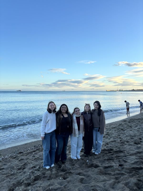 Five study abroad students standing together on a beach in front of a body of water and smiling at the camera