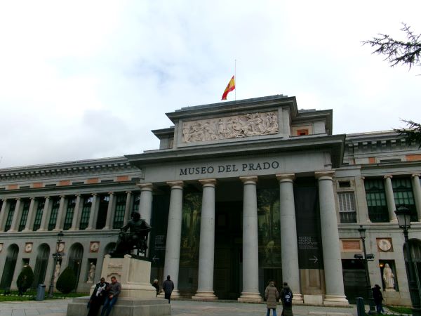 A large concrete building with a statue and people standing in front and a Spanish flag flying from the top