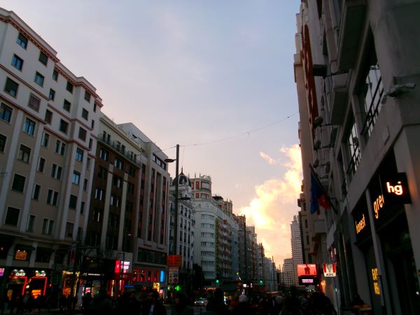 A city street during evening, with low natural light, bright clouds in the distance, and store lights light up at street level