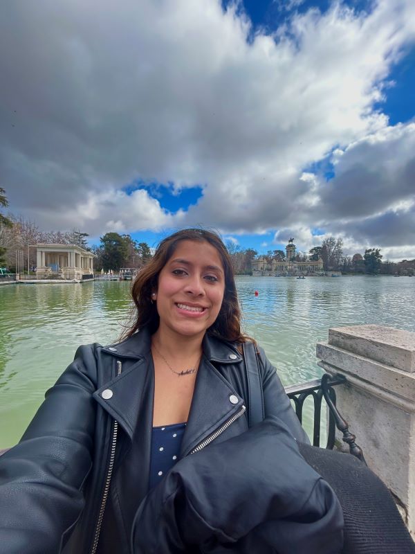 A study abroad student taking a selfie in front of a large pond with large bright clouds in the distant sky