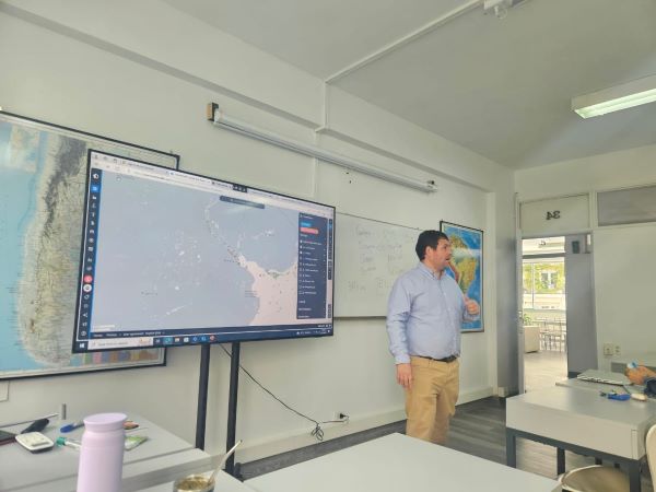A person standing at the front of a classroom next to a TV screen featuring a blue and white map