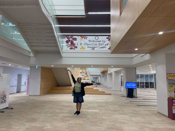 A study abroad student standing in the interior of a large bright building, smiling at the camera with their arms held wide to the sides