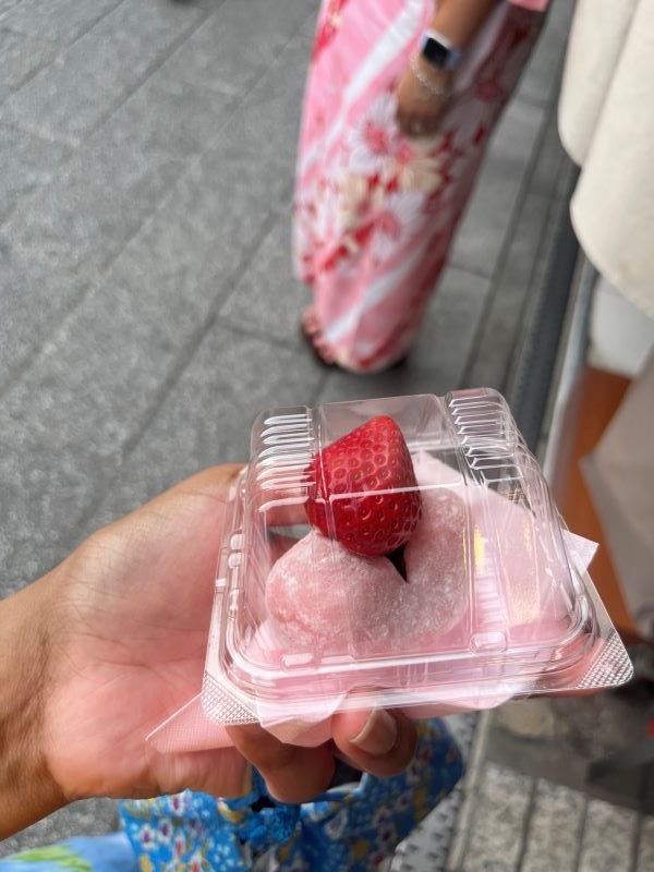 A close-up of a hand holding a small plastic container containing a small pink dessert bun with a bright red strawberry atop it