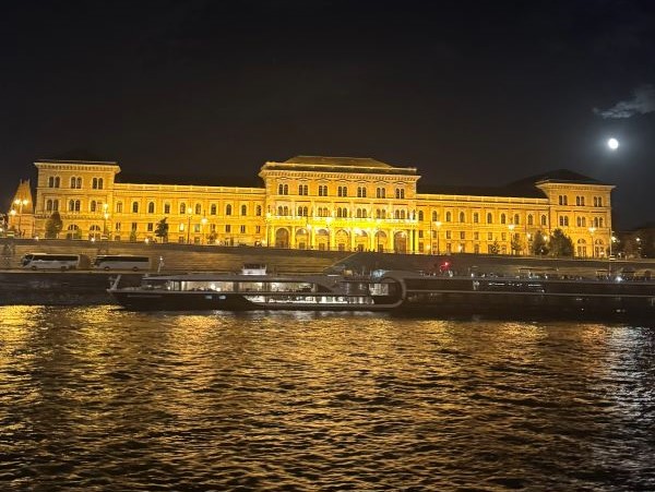 A large ornate building seen across a body of water and bathed in golden light during nighttime with a full moon in the sky above it