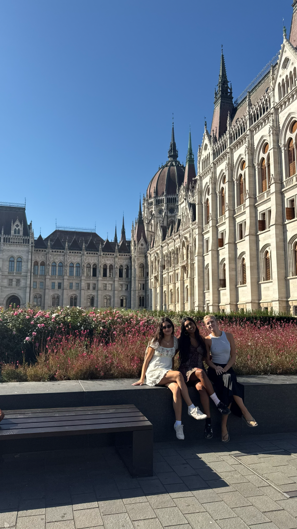 Three study abroad students sitting on a bench outside of a large ornate building and smiling at the camera