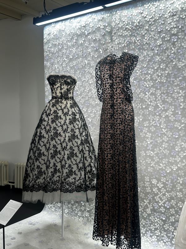 Two intricately-crafted black dresses on display in front of a lacy screen in an indoor setting
