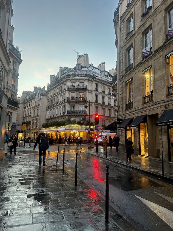 A Parisian street after rainfall under an evening sky, with bright lights from stores casting reflections on the wet pavement