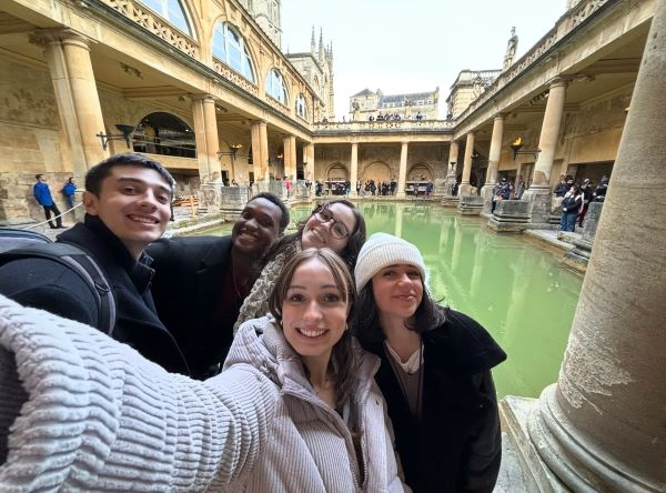 Five study abroad students taking a selfie in front of a large ancient green-colored indoor Roman bath