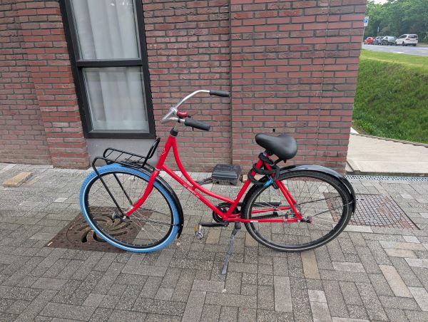 A red bicycle with a light blue front wheel standing outdoors in front of a building