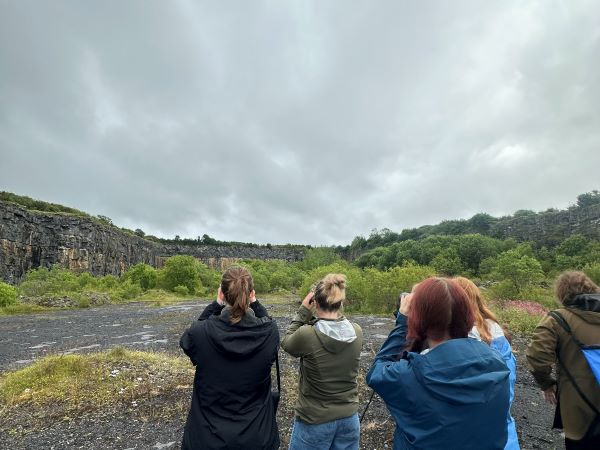 A group of five study abroad students facing away from the camera and pointing binoculars toward a cloudy gray sky