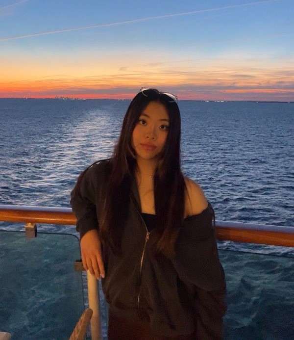 A study abroad student with long dark hair leaning on a railing in front of a large body of water and smiling at the camera