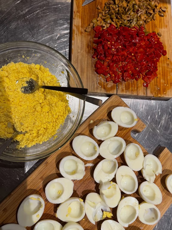 A steel tabletop featuring a wooden cutting boards with chopped eggs, peppers, and olives