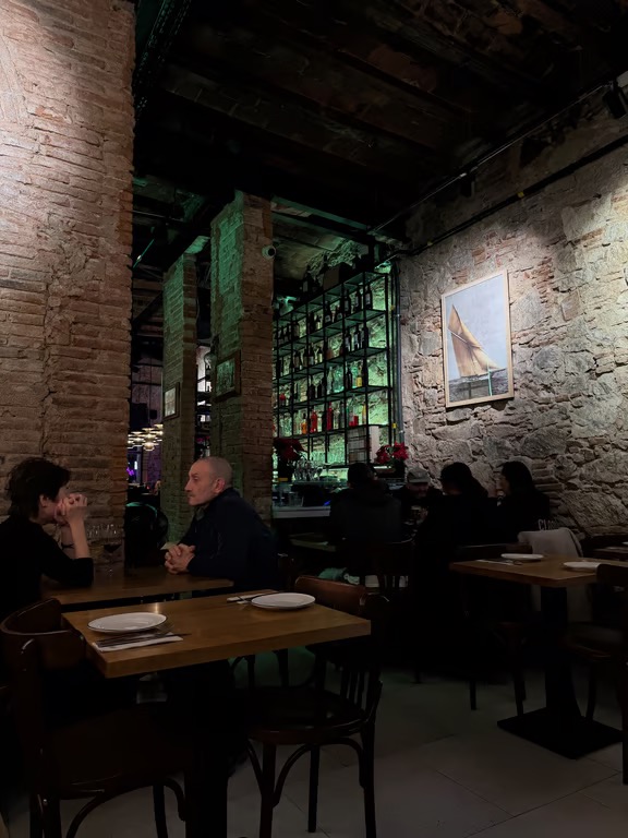A dimly-lit restaurant interior, featuring stone walls and people sitting at small wooden tables