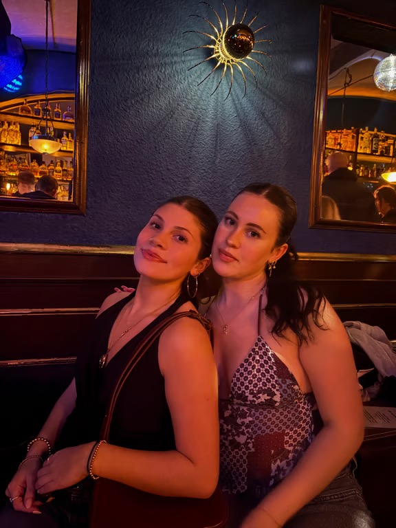 A study abroad student slightly leaning on their friend, inside a dimly-lit interior space, both looking at the camera