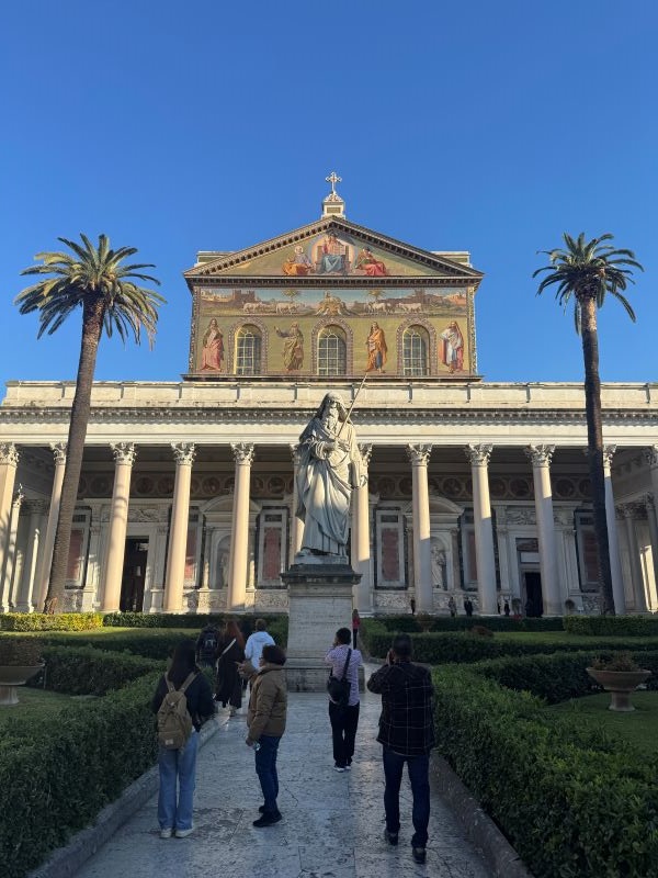 People standing in front of a tall stone coloannaded building with a large statue of a person in front of it and palm trees on either side of the statue
