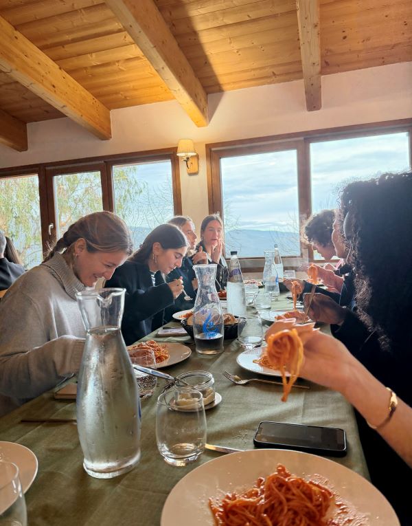 A group of study abroad students sat at a table indoors, smiling, and eating plates of spaghetti