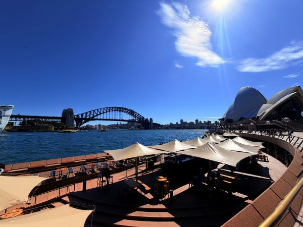 A large building seen to the right with covered tables in the foreground and a body of water and large bridge to the left, all under a bright blue sky