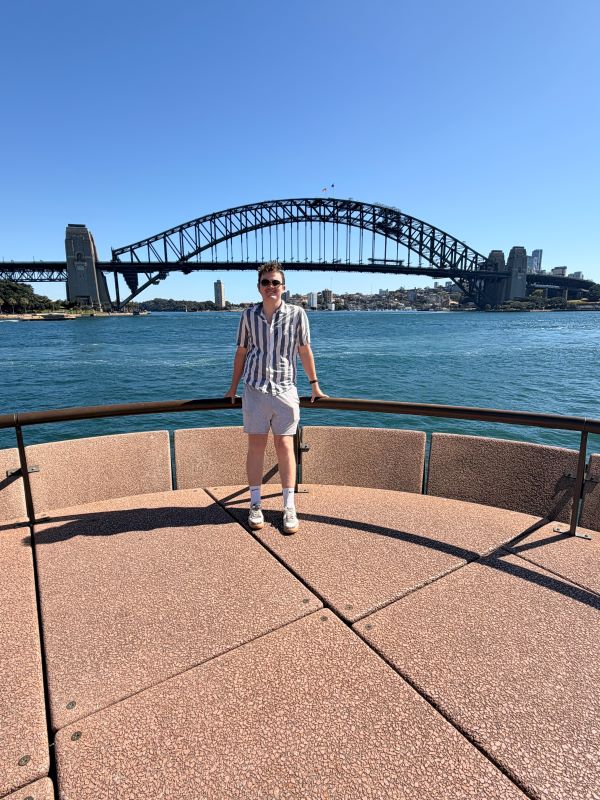 A study abroad student wearing sunglasses and smiling at the camera while standing outdoors in front of a large bridge over a body of water