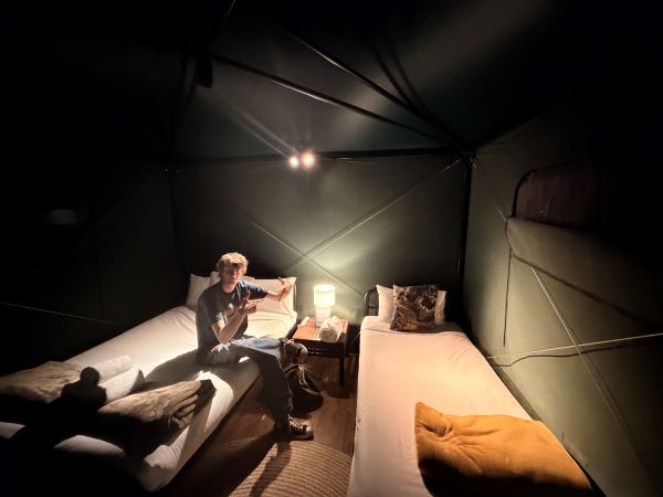 A study abroad student sitting on a bed in an indoor space and smiling at the camera while holding their hands up in peace signs