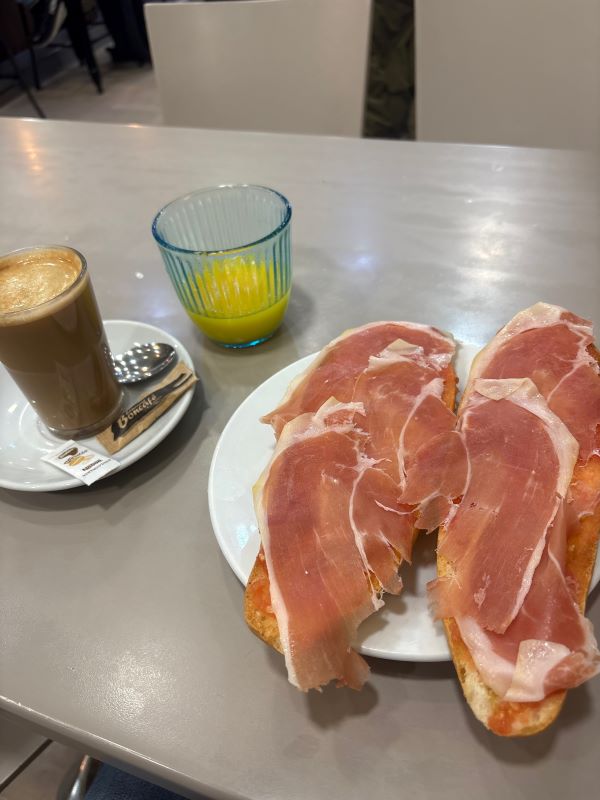 A plate with toast covered in thin slices of ham sat next to a plate with a cup of coffee on it and a glass juice with a small amount of orange juice