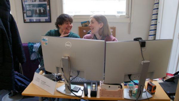 Two people sitting at a desk between two large computer monitors, smiling and looking at each other